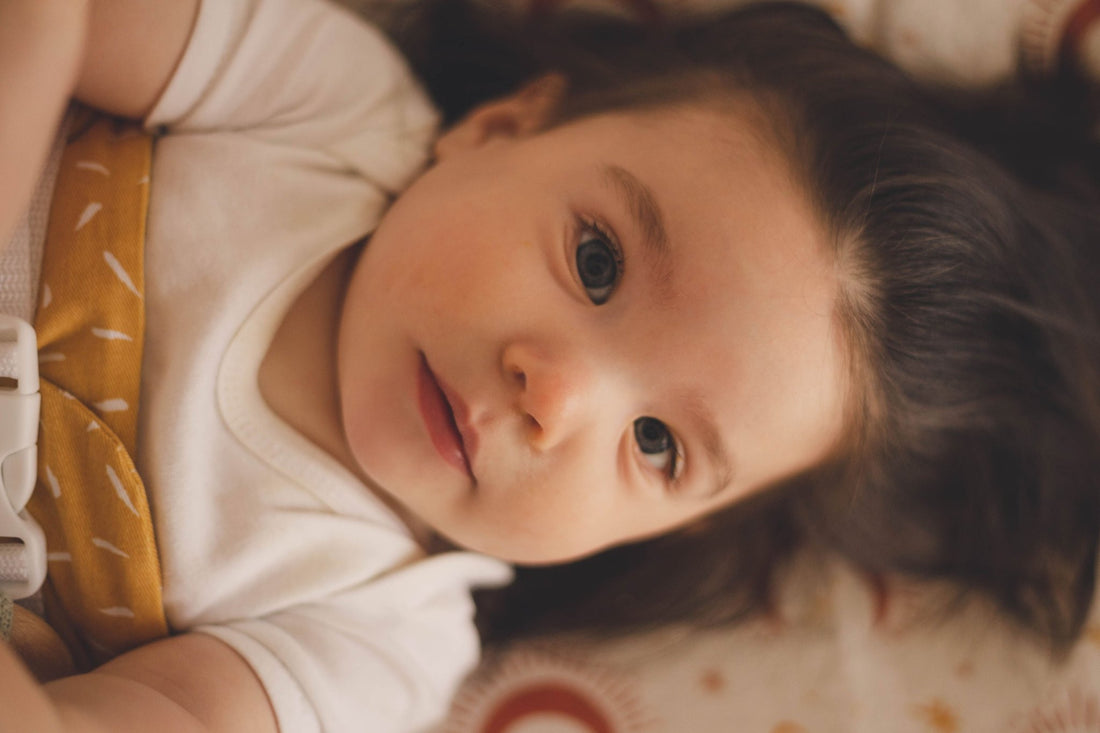 A close up of a baby with brown hair, looking at the camera. She is wrapped in a Butterscotch Sprinkle WriggleBum nappy change harness