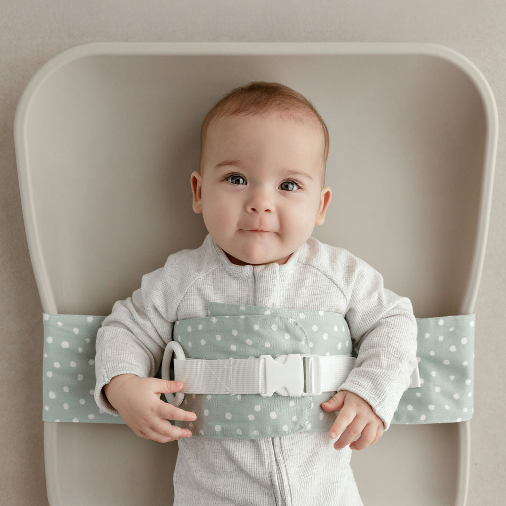 Wriggly baby in sage green WriggleBum nappy change harness, lying on a Leander Matty change mat