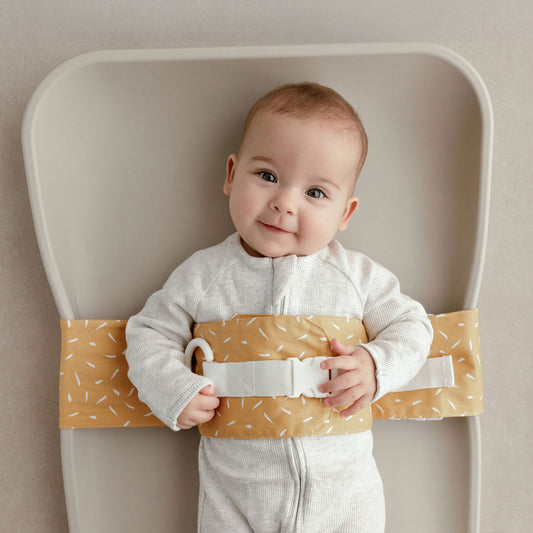 Wriggly smiling baby in mustard Butterscotch Sprinkle WriggleBum nappy change harness, lying on a Leander Matty change mat