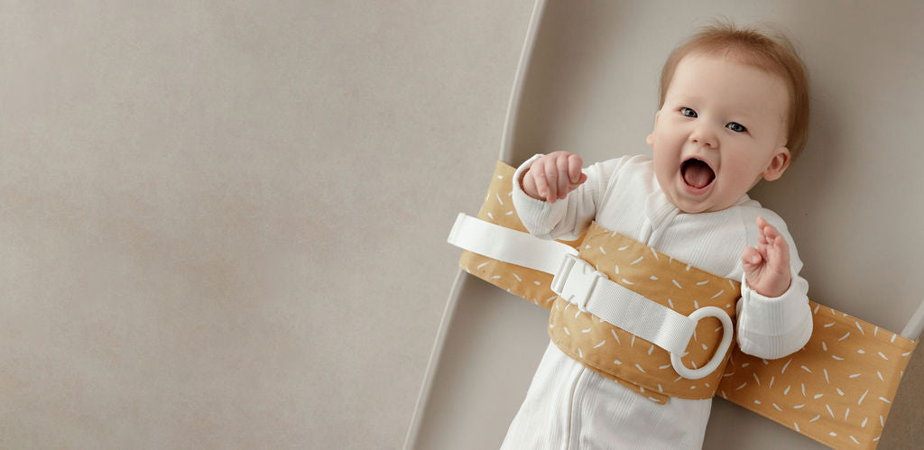 Wriggly baby in a yellow and white WriggleBum nappy change harness on a Leander Matty change mat