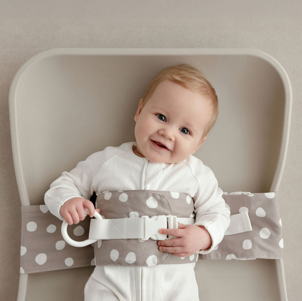 Wriggly smiling baby in grey and white Spotty Dotty WriggleBum nappy change harness, lying on a Leander Matty change mat
