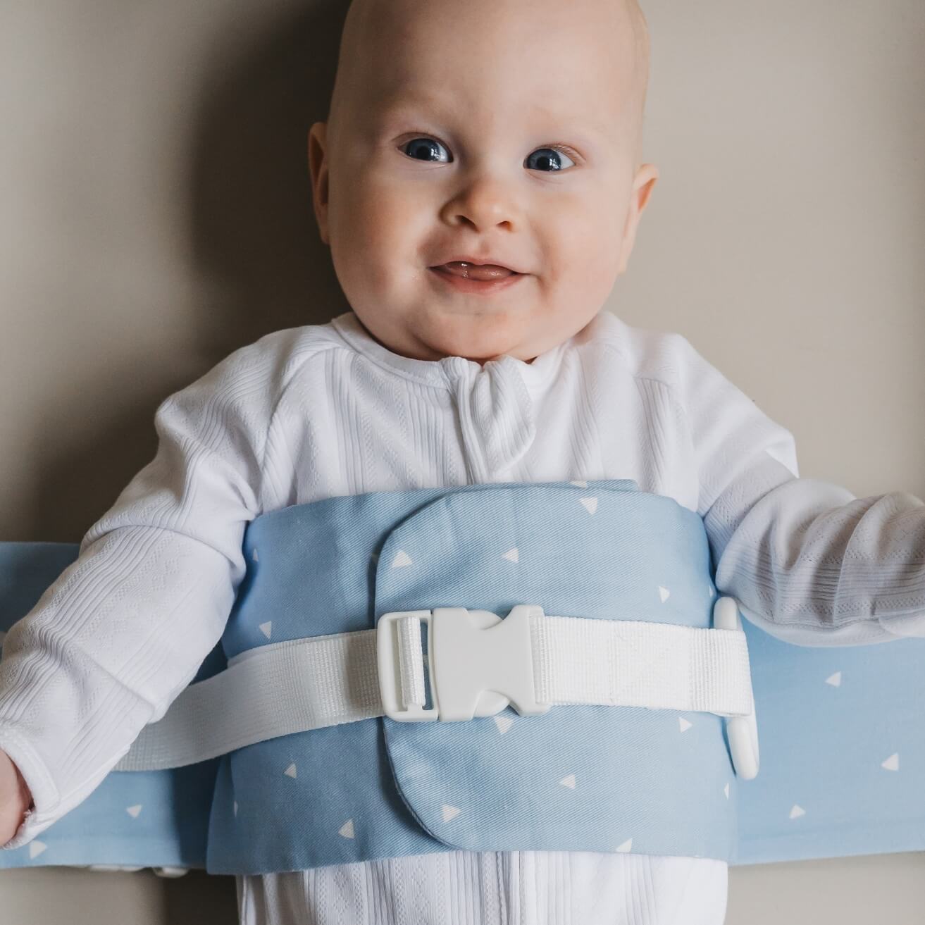 A baby is lying on a solid change mat, wrapped in a Breezy Blue WriggleBum nappy change harness. The baby is smiling.