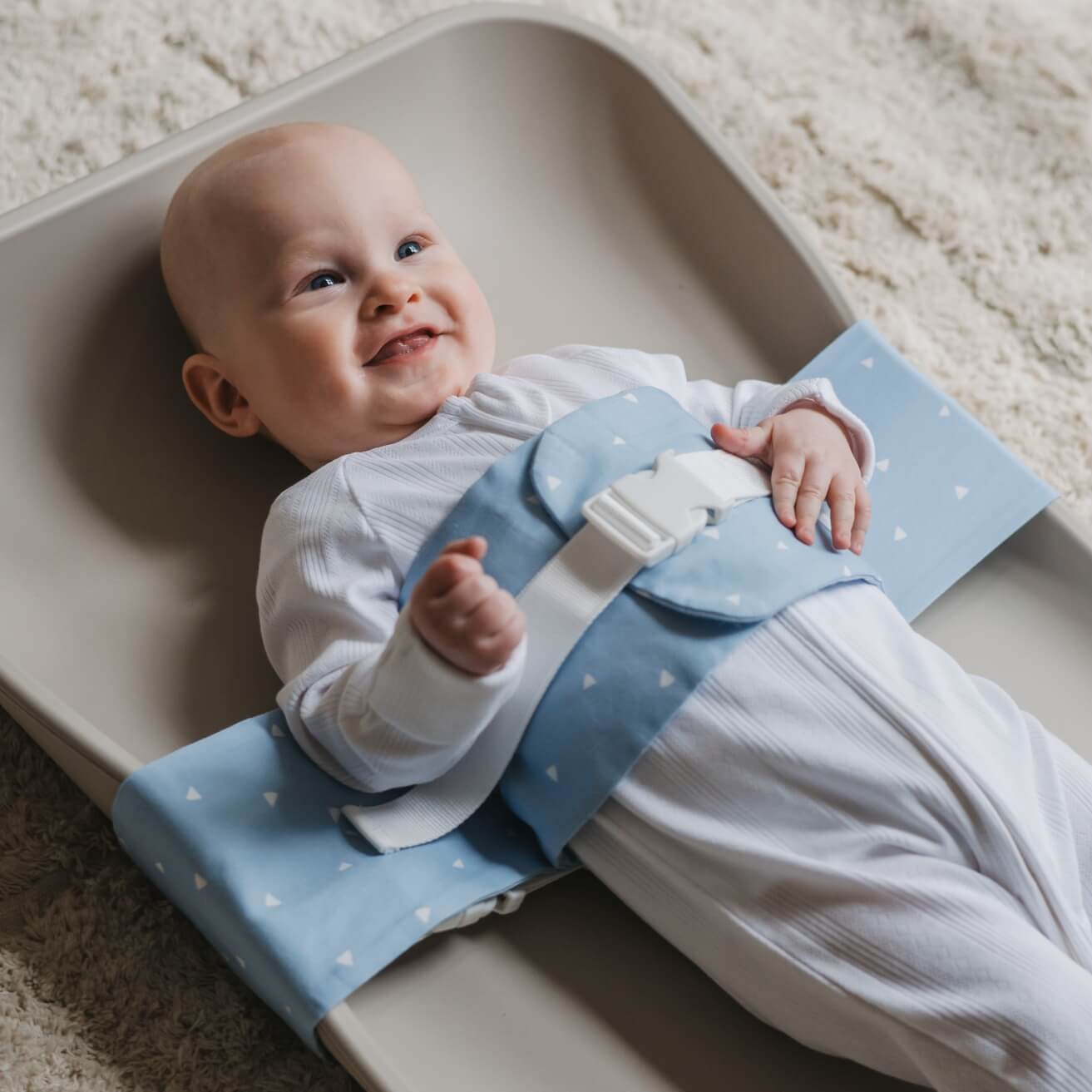 A baby is lying on a solid change mat, wrapped in a Breezy Blue WriggleBum nappy change harness. The baby is smiling.