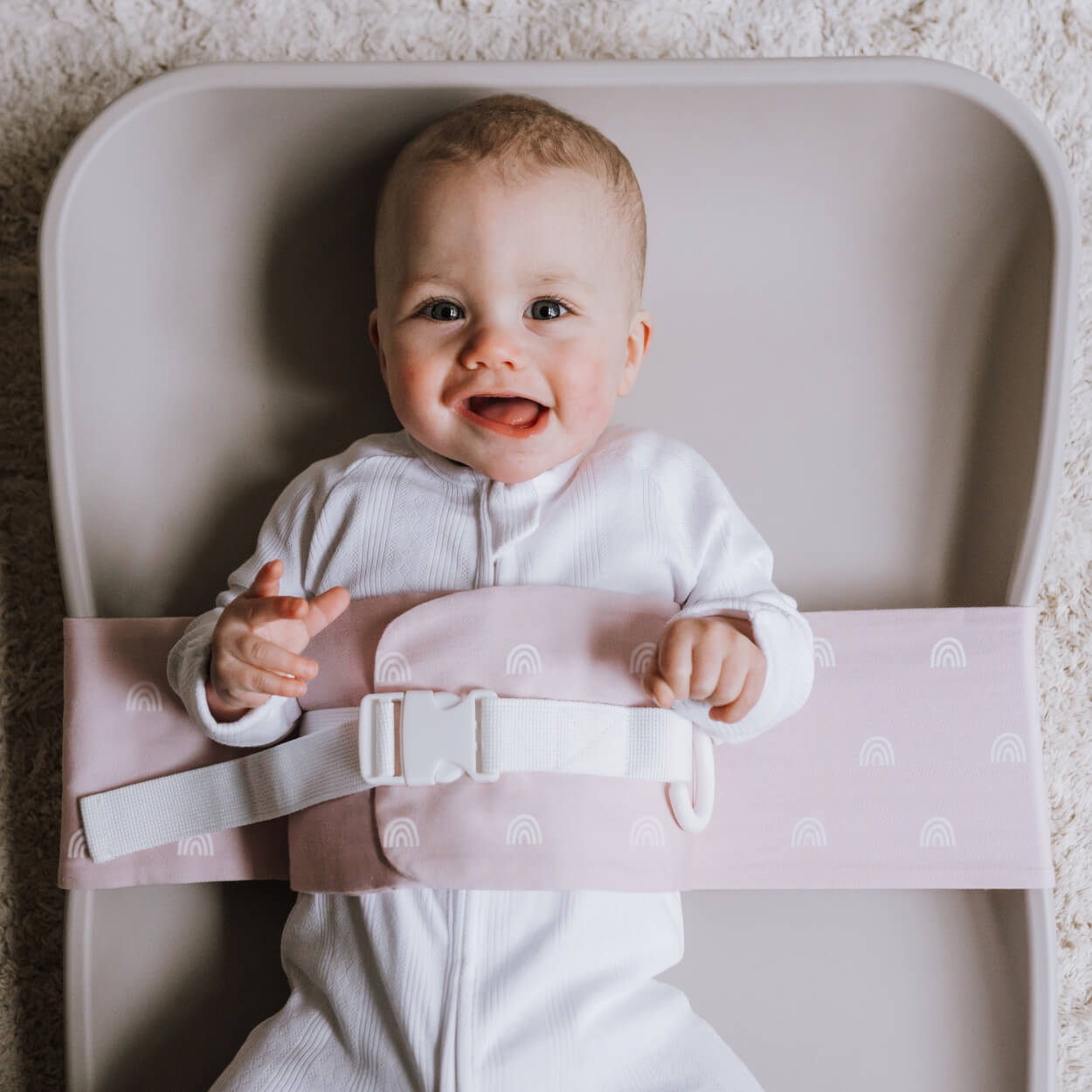 A baby is lying on a solid change mat, wrapped in an Rainbow Blush WriggleBum nappy change harness. He is looking at the camera and smiling.