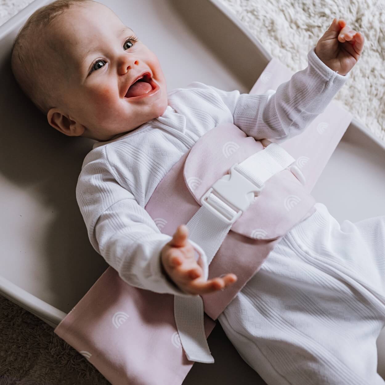 A baby is lying on a solid change mat, wrapped in an Rainbow Blush WriggleBum nappy change harness. He is smiling.