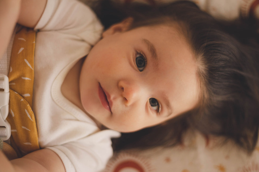 A baby with dark brown hair is lying on a change table and staring at camera. They are wrapped in a WriggleBum nappy change harness.