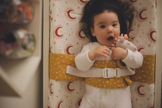 A baby with dark brown hair is lying on a change table and wrapped in a Butterscotch Sprinkle WriggleBum nappy change harness. She is playing with a toy attached to the harness.