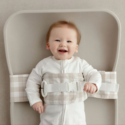 Wriggly smiling baby in beige WriggleBum nappy change harness, lying on a Leander Matty change mat