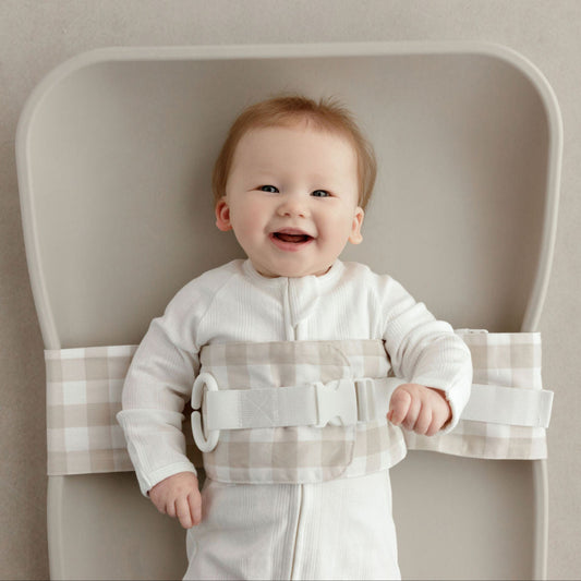Wriggly smiling baby in beige WriggleBum nappy change harness, lying on a Leander Matty change mat