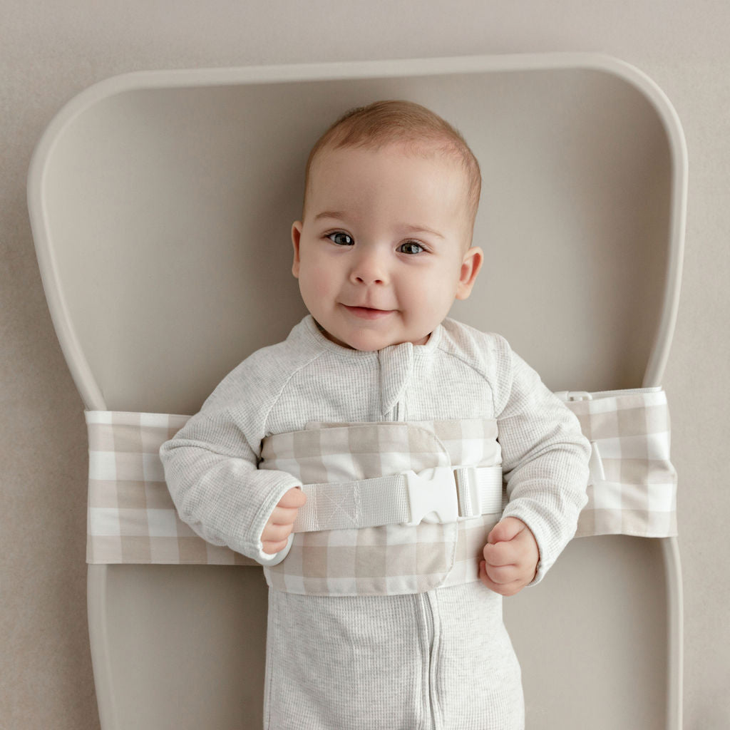 Wriggly smiling baby in beige WriggleBum nappy change harness, lying on a Leander Matty change mat