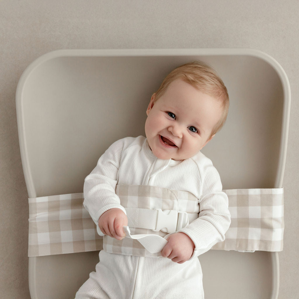Wriggly smiling baby in beige WriggleBum nappy change harness, lying on a Leander Matty change mat