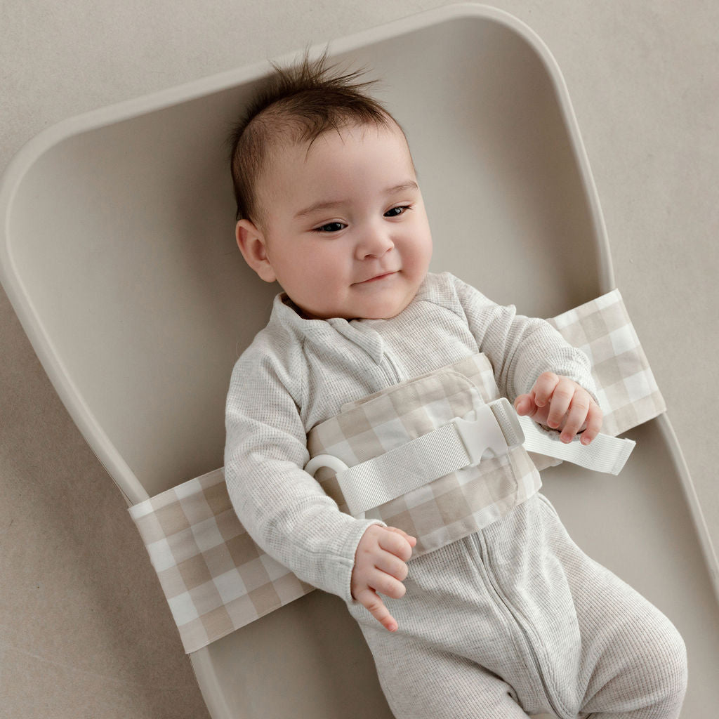 Wriggly smiling baby in beige WriggleBum nappy change harness, lying on a Leander Matty change mat