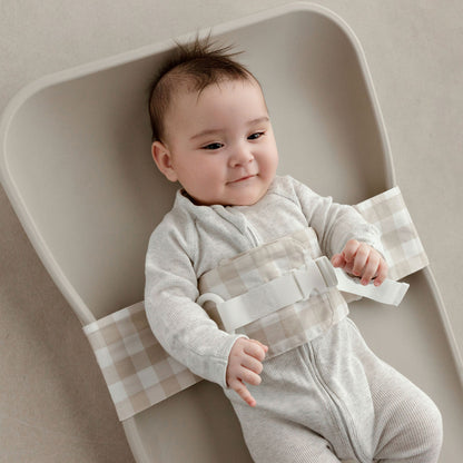 Wriggly smiling baby in beige WriggleBum nappy change harness, lying on a Leander Matty change mat