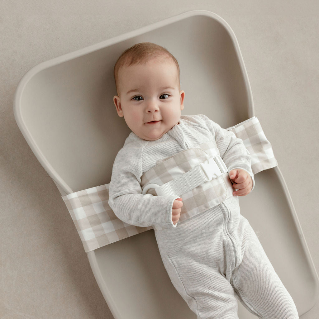 Wriggly smiling baby in beige WriggleBum nappy change harness, lying on a Leander Matty change mat
