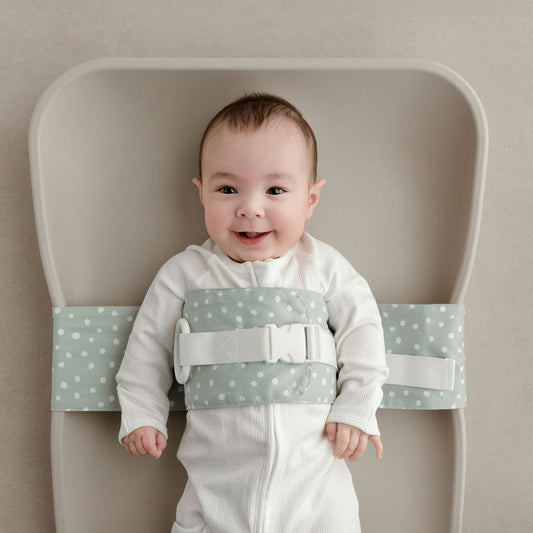 Wriggly smiling baby in sage green WriggleBum nappy change harness, lying on a Leander Matty change mat