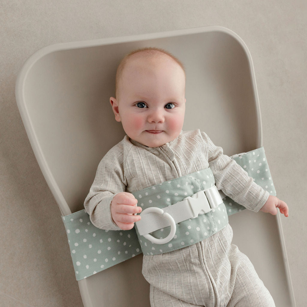 Wriggly baby in sage green WriggleBum nappy change harness, lying on a Leander Matty change mat