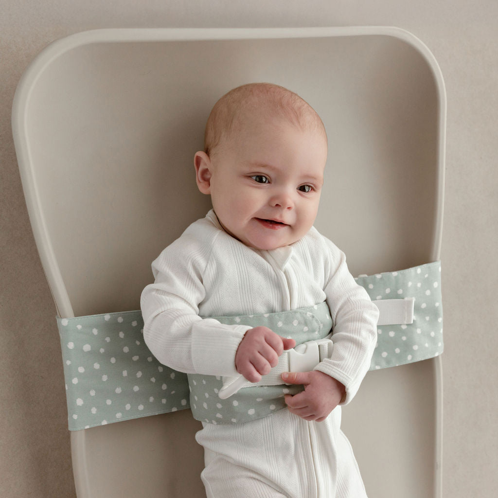 Wriggly smiling baby in sage green WriggleBum nappy change harness, lying on a Leander Matty change mat