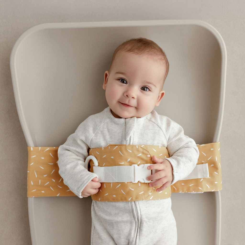 Wriggly smiling baby in mustard Butterscotch Sprinkle WriggleBum nappy change harness, lying on a Leander Matty change mat
