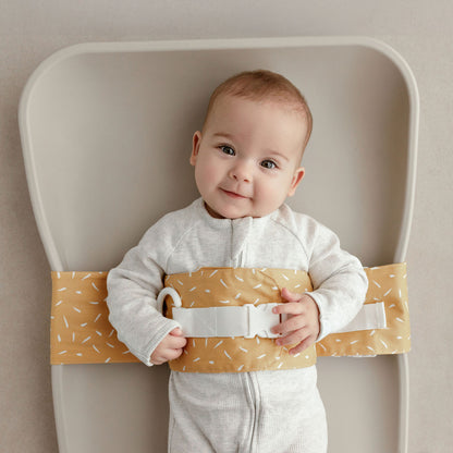 Wriggly smiling baby in mustard Butterscotch Sprinkle WriggleBum nappy change harness, lying on a Leander Matty change mat
