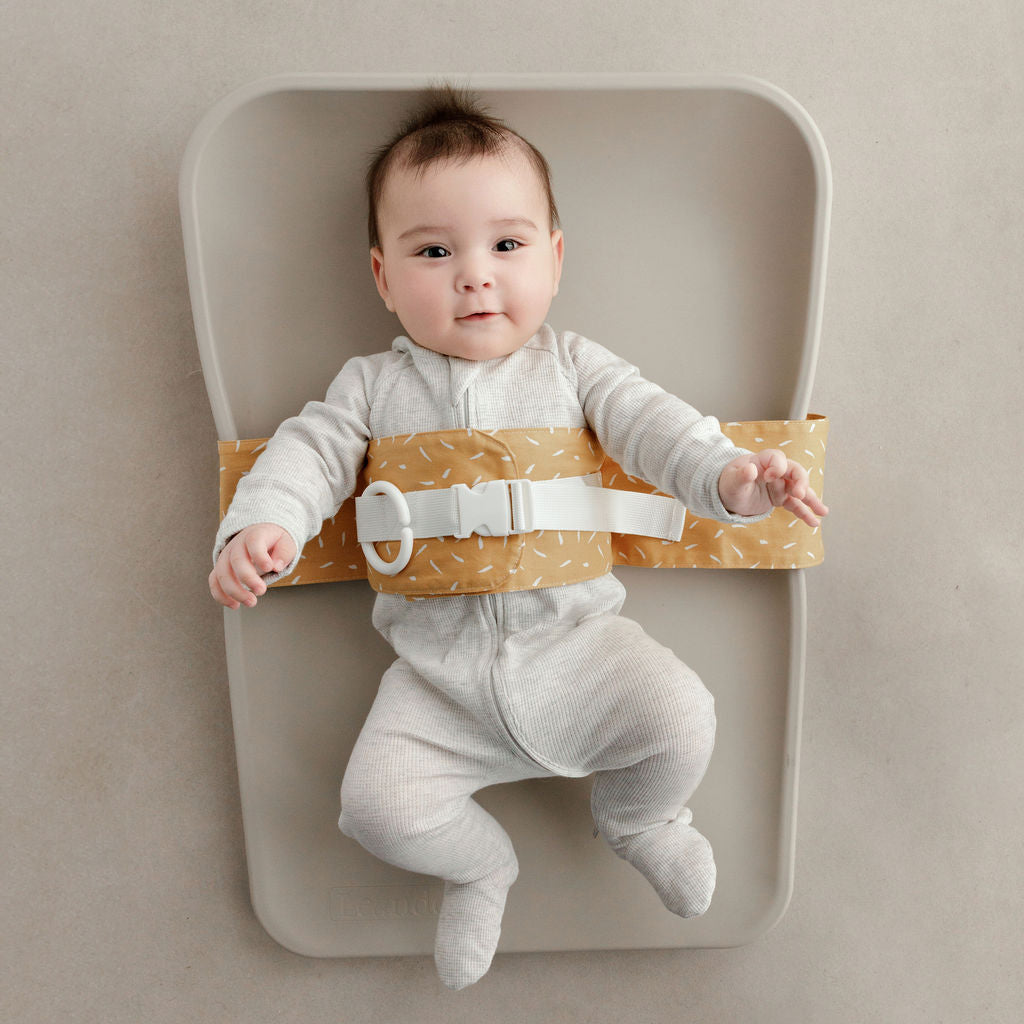 Wriggly smiling baby in mustard Butterscotch Sprinkle WriggleBum nappy change harness, lying on a Leander Matty change mat.