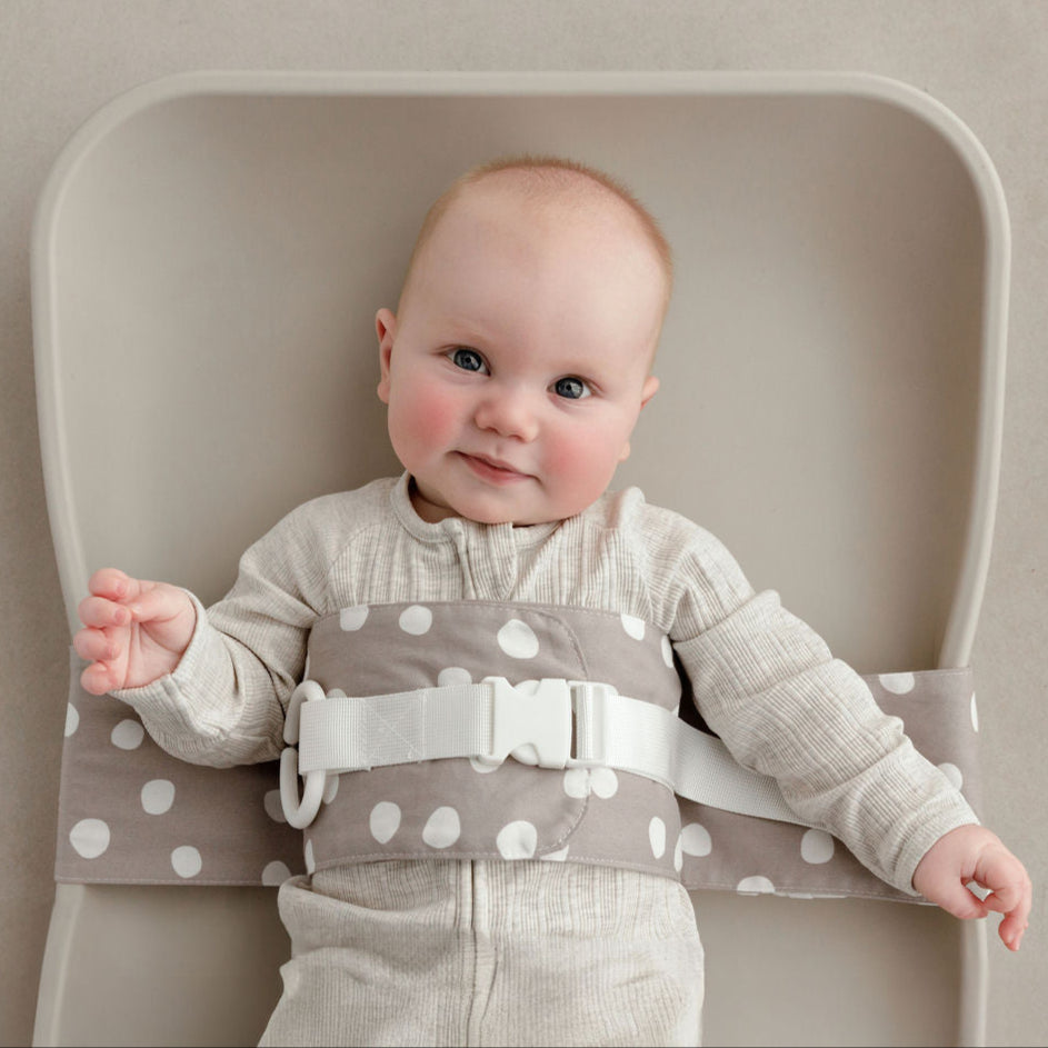 Wriggly smiling baby in grey and white Spotty Dotty WriggleBum nappy change harness, lying on a Leander Matty change mat 