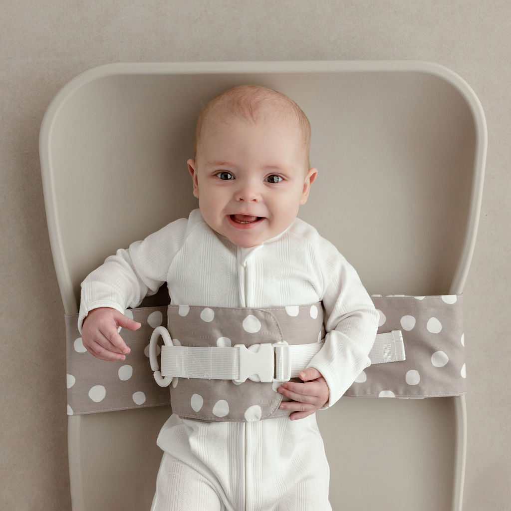 Wriggly smiling baby in grey and white Spotty Dotty WriggleBum nappy change harness, lying on a Leander Matty change mat 