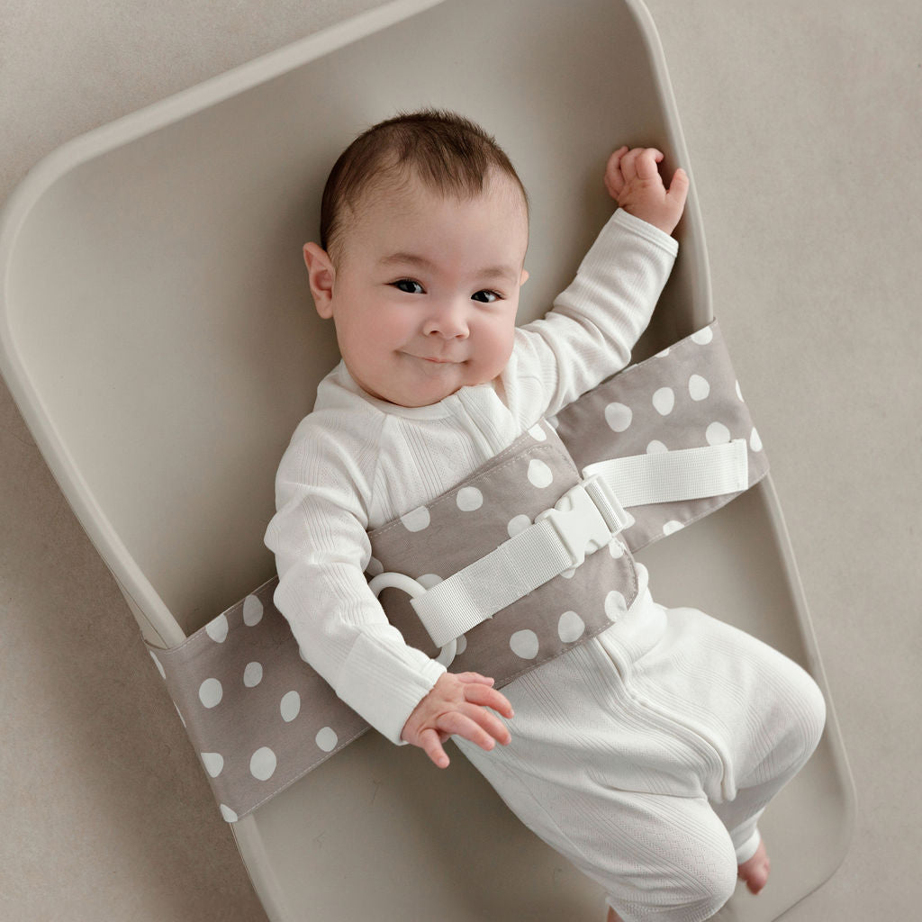 Wriggly smiling baby in grey and white Spotty Dotty WriggleBum nappy change harness, lying on a Leander Matty change mat 