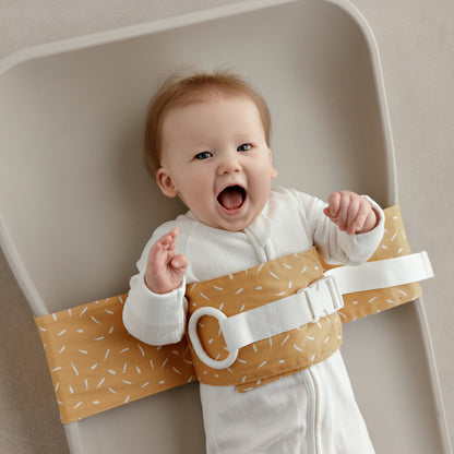 Smiling baby lying on a change mat, with a Butterscotch Sprinkle WriggleBum nappy change harness wrapped around them. 
