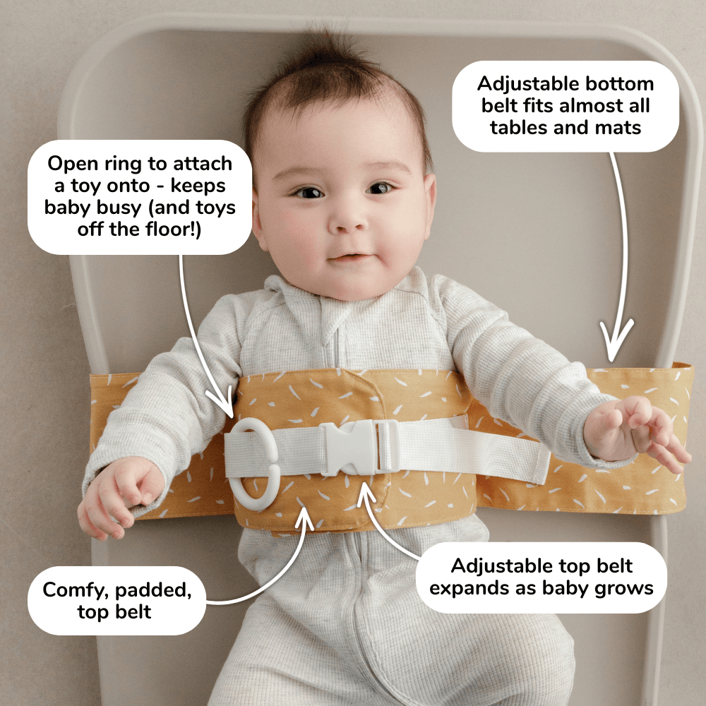 Baby lies on a change mat with an Butterscotch Sprinkle WriggleBum nappy change harness wrapped around them. 
Four white bubbles highlight the key features of the harness: 
- Open ring to attach a toy onto - keeps baby busy (and toys off the floor!)
- Adjustable bottom belt fits almost all tables and mats 
- Adjustable top belt expands as baby grows
- Comfy, padded top belt