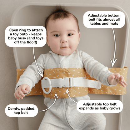Baby lies on a change mat with an Butterscotch Sprinkle WriggleBum nappy change harness wrapped around them. 
Four white bubbles highlight the key features of the harness: 
- Open ring to attach a toy onto - keeps baby busy (and toys off the floor!)
- Adjustable bottom belt fits almost all tables and mats 
- Adjustable top belt expands as baby grows
- Comfy, padded top belt