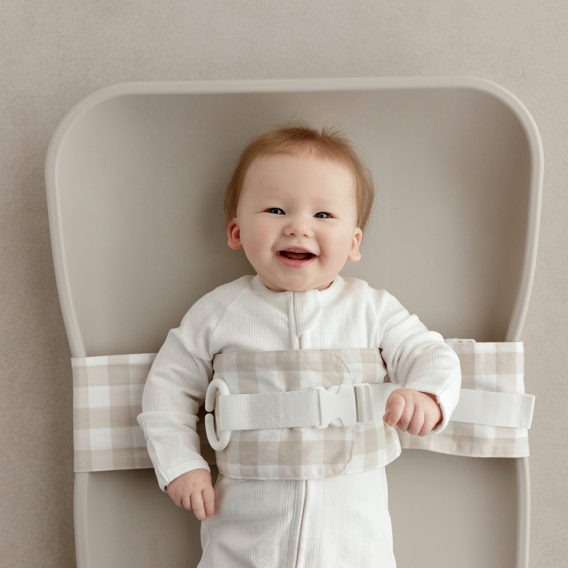 Smiling baby lying on a change mat, with a Oatmeal Afternoon WriggleBum nappy change harness wrapped around them. 
