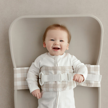 Smiling baby lying on a change mat, with a Oatmeal Afternoon WriggleBum nappy change harness wrapped around them. 
