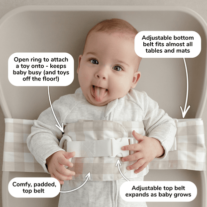 Baby lies on a change mat with an Oatmeal Afternoon WriggleBum nappy change harness wrapped around them. 
Four white bubbles highlight the key features of the harness: 
- Open ring to attach a toy onto - keeps baby busy (and toys off the floor!)
- Adjustable bottom belt fits almost all tables and mats 
- Adjustable top belt expands as baby grows
- Comfy, padded top belt