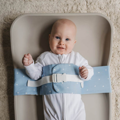 A baby is lying on a solid change mat, wrapped in a Breezy Blue WriggleBum nappy change harness. The baby is smiling. 