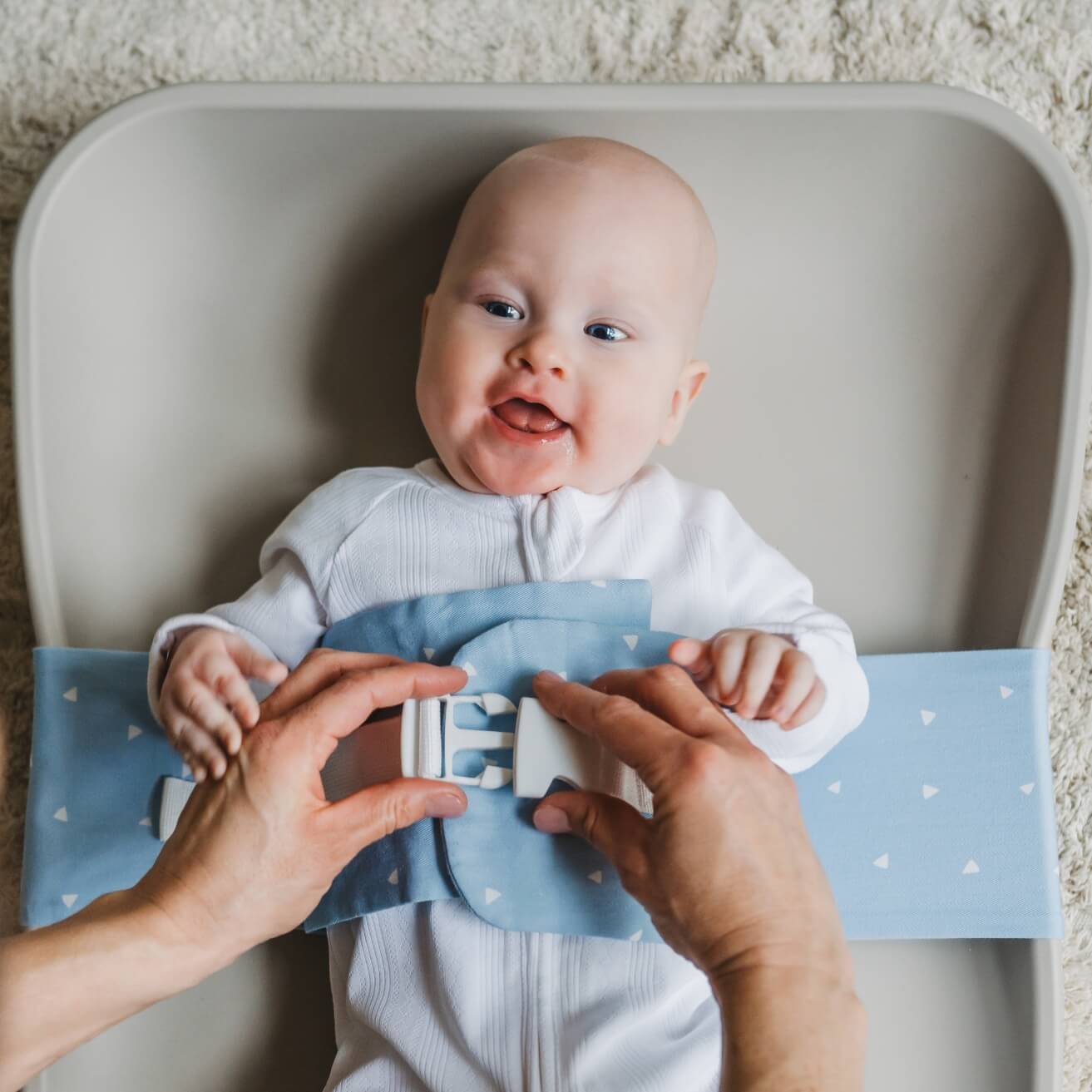 A baby is lying on a solid change mat, wrapped in a Breezy Blue WriggleBum nappy change harness. An adult, who is out of frame, is clipping the buckle on the harness.