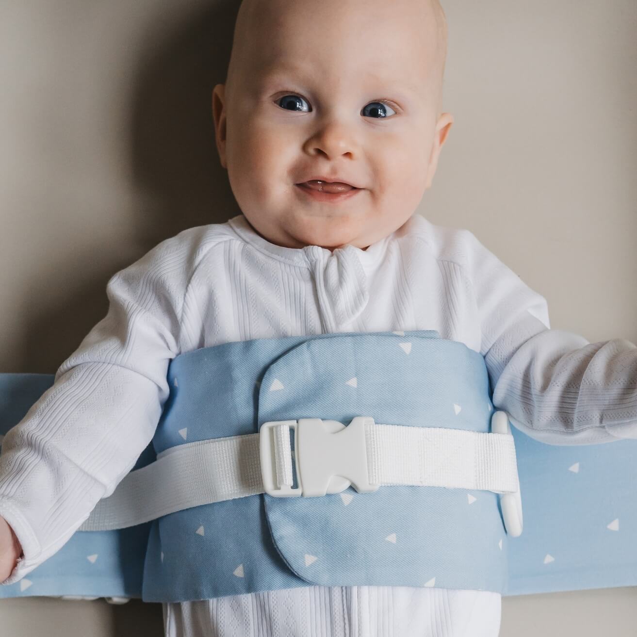 A baby is lying on a solid change mat, wrapped in a Breezy Blue WriggleBum nappy change harness. The baby is smiling. 