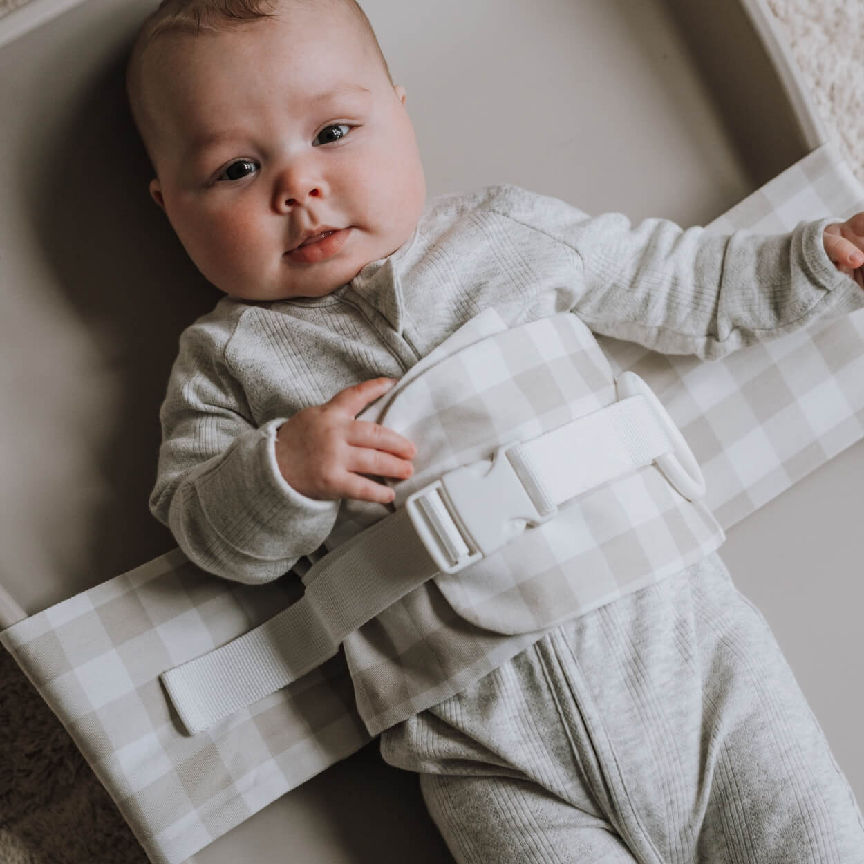 A baby is lying on a solid change mat, wrapped in an Oatmeal Afternoon WriggleBum nappy change harness. She is looking at the camera.