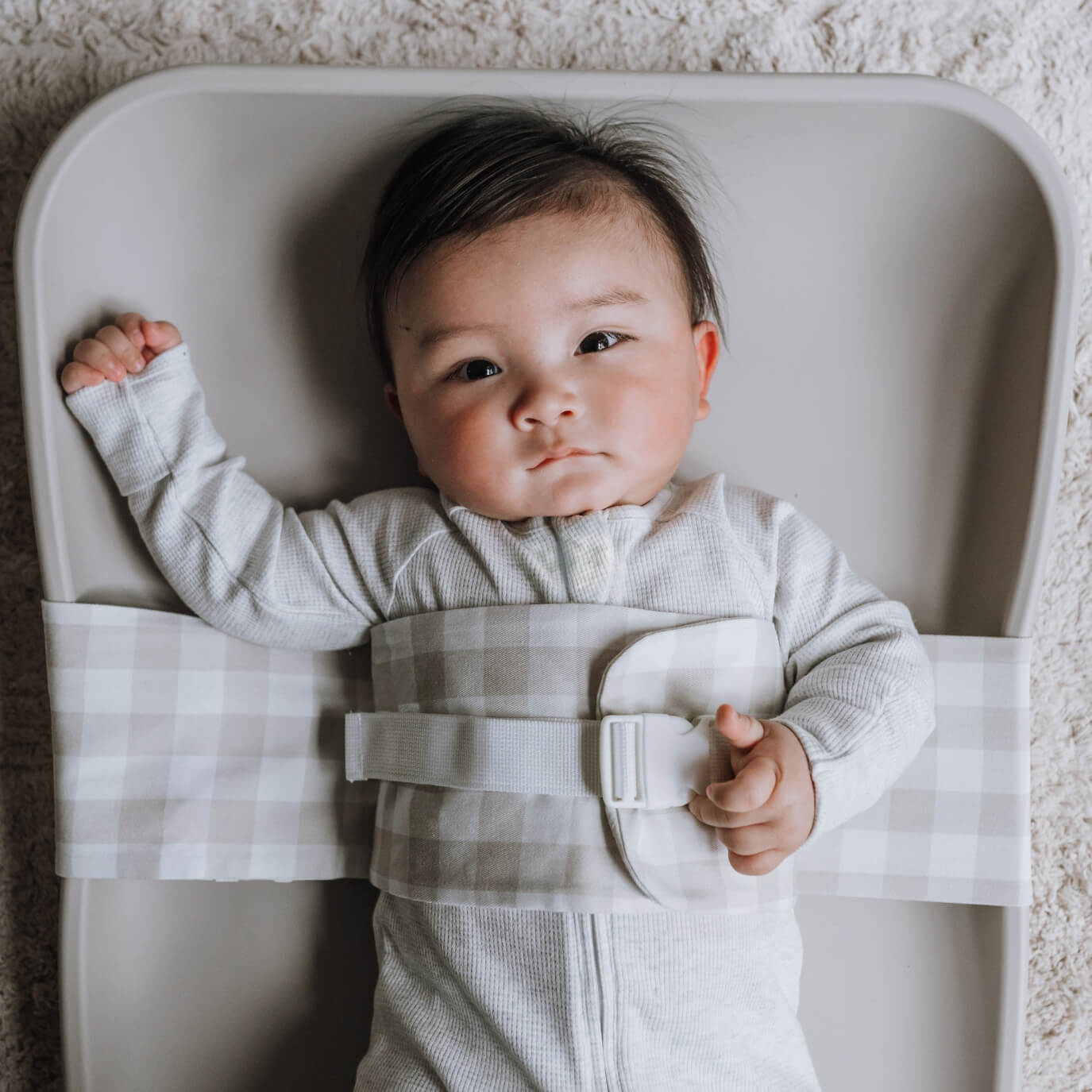 A baby is lying on a solid change mat, wrapped in an Oatmeal Afternoon WriggleBum nappy change harness. He is looking at the camera.