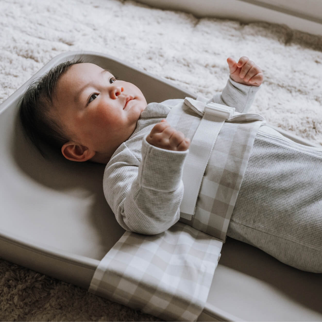 A baby is lying on a solid change mat, wrapped in an Oatmeal Afternoon WriggleBum nappy change harness.