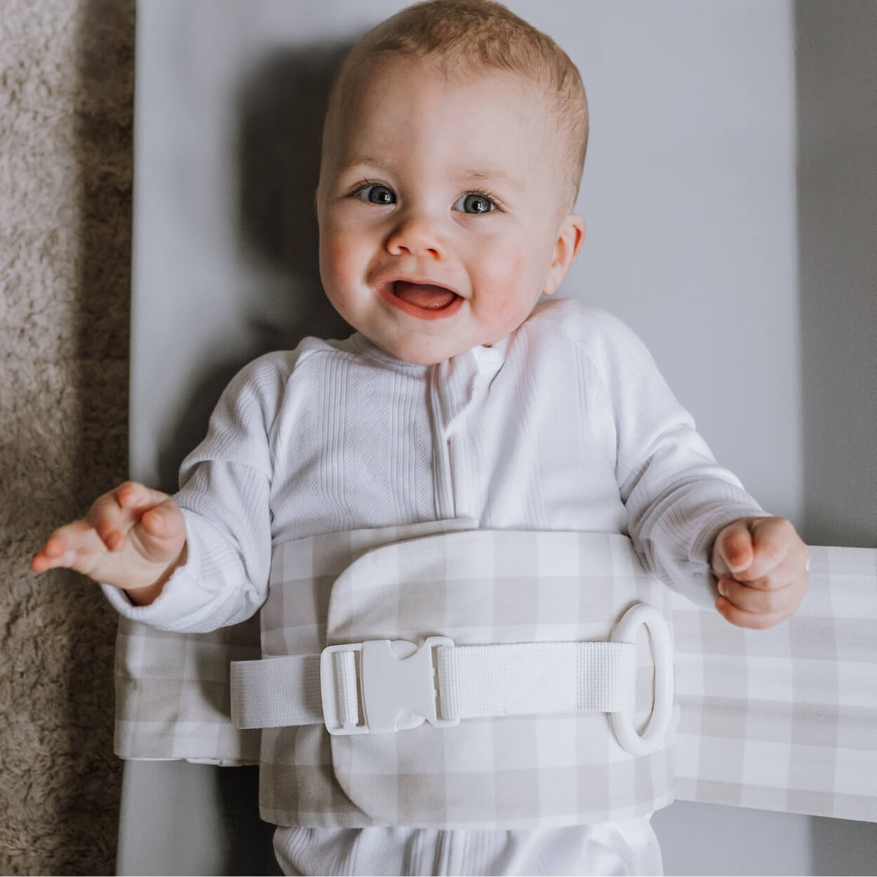 A baby is lying on a solid change mat, wrapped in an Oatmeal Afternoon WriggleBum nappy change harness. He is looking at the camera and smiling.