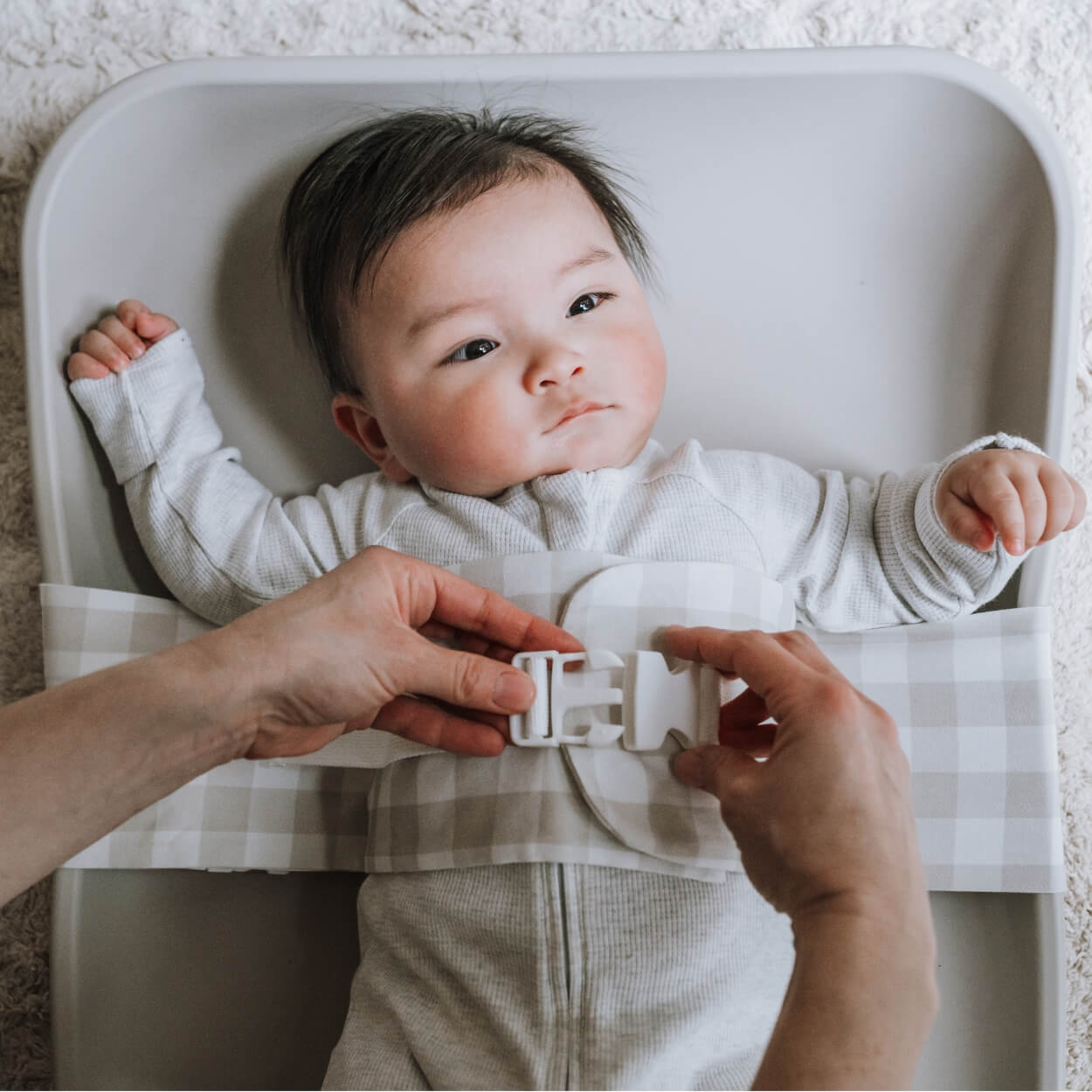 A baby is lying on a solid change mat, wrapped in an Oatmeal Afternoon WriggleBum nappy change harness. An adult, who is out of frame, is clipping the buckle on the harness. 