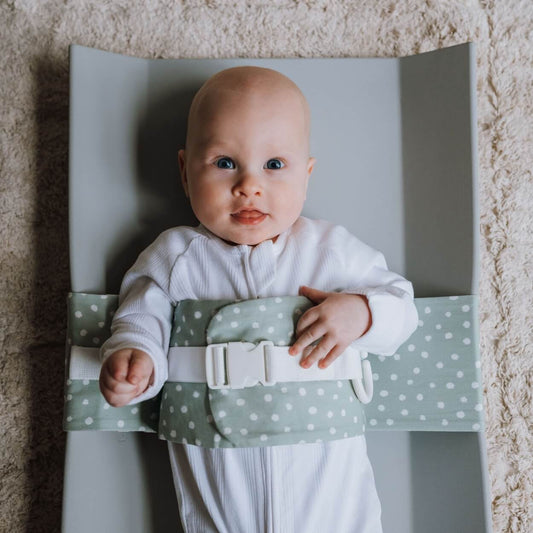 A blue eyed baby is lying on a solid change mat, wrapped in a Sage Whimsy WriggleBum nappy change harness. He is staring at the camera.